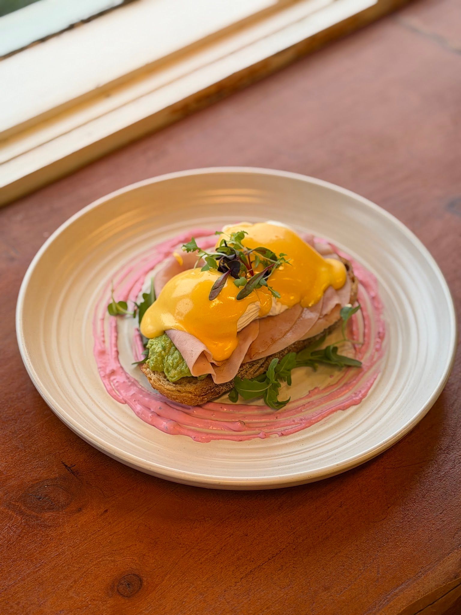 Eggs benedict with hollandaise, shaved ham and avocado on pink beetroot cream in a ceramic bowl