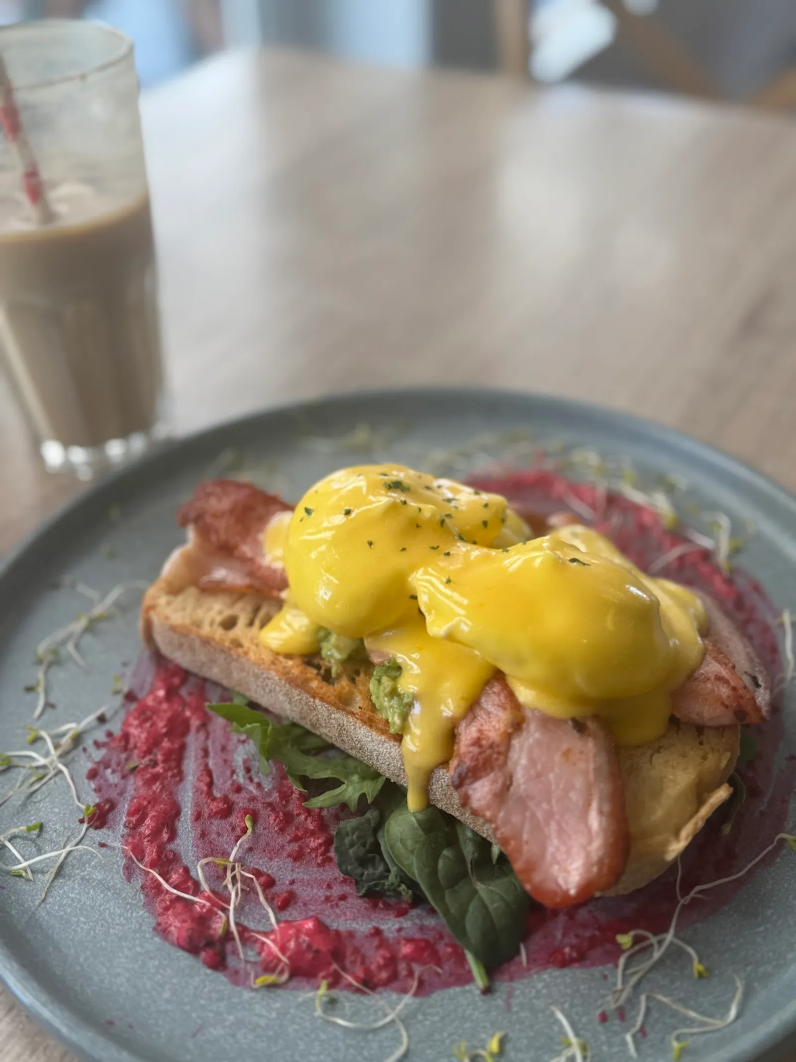 Eggs benedict with hollandaise over bacon and spinach on pink beetroot hummus, with milkshake in background