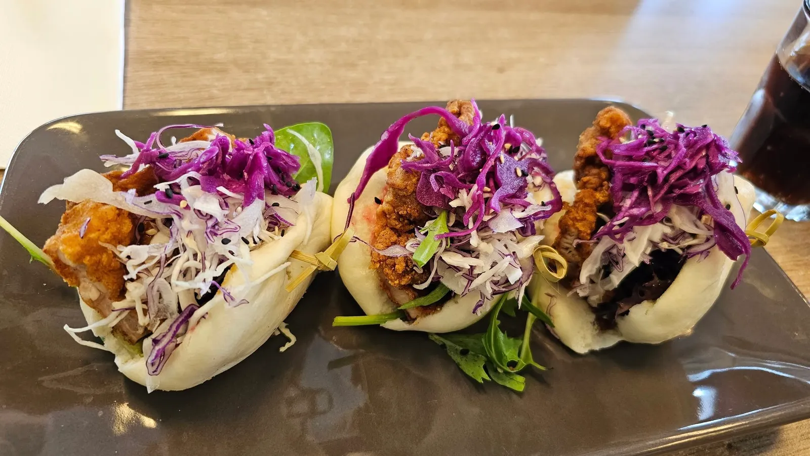 Three steamed bao buns with crispy chicken and purple cabbage slaw on a dark ceramic plate