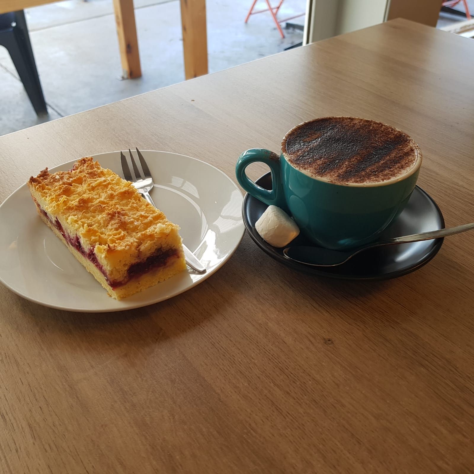 Berry crumble slice on saucer beside a flat white cappuccino in a teal cup on a timber table