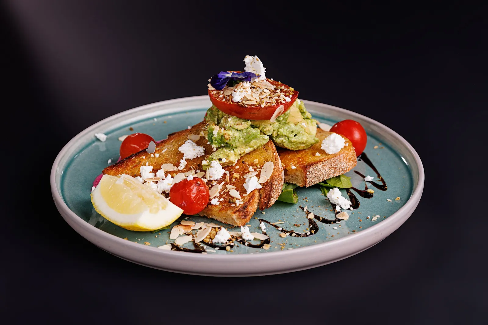 Gourmet avocado toast with roasted tomatoes, feta, almonds and edible flowers on black background