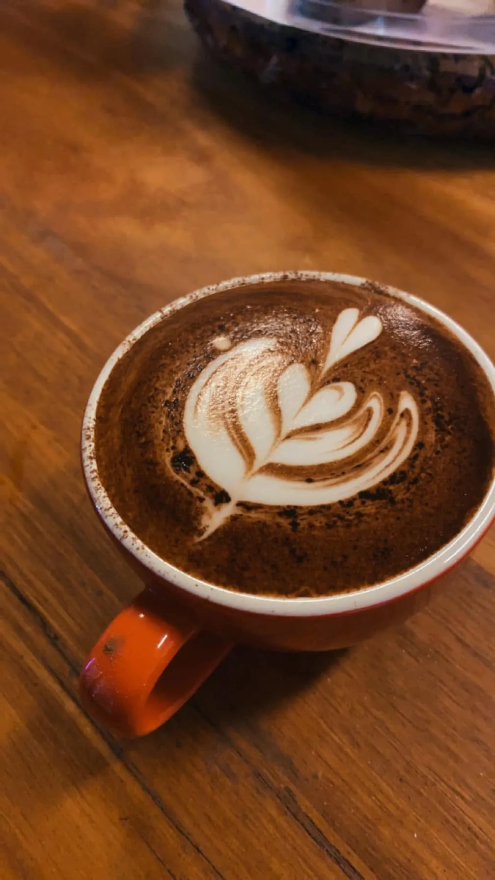 Mocha with latte art leaf in an orange ceramic mug on a timber cafe table